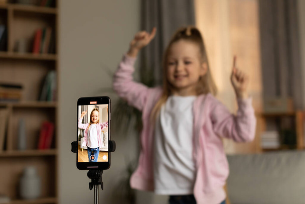 Kid Blogging. Cheerful Blogger Girl Making Video On Cellphone Dancing Posing At Home. Child Filming Blog And Having Fun In Modern Living Room. Selective Focus On Mobile Phone - Foto, Imagen