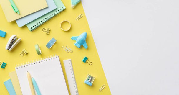 School accessories concept. Top view panoramic photo of colorful stationery plane shaped sharpener notepads ruler pens binder clips adhesive tape and stapler on bicolor yellow and white background - Fotó, kép