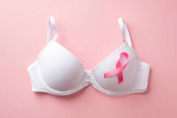 Breast cancer awareness concept. Top view photo of white brassiere with pink silk ribbon on isolated pastel pink background - Φωτογραφία, εικόνα
