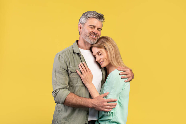 Portrait of loving middle aged spouses hugging with closed eyes while standing isolated over yellow studio background. Romantic mature couple expressing their love with each other - Photo, image