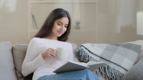 Serene young girl student daughter relaxed arabian biracial intelligent woman sits on sofa reads paper book scientific literature educational textbook preparing for homework exams enjoying bestseller - Imágenes, Vídeo