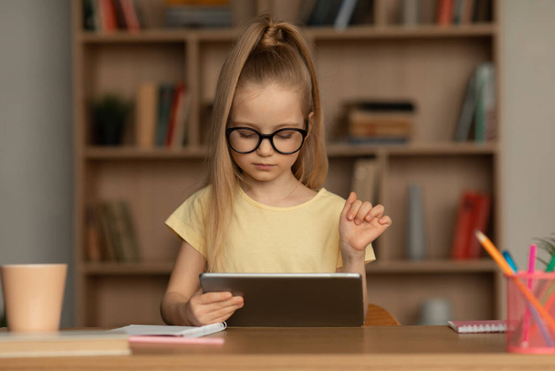 School Kid Girl met behulp van digitale tablet doet huiswerk Online zitten aan de balie thuis. Kinderen leren online met behulp van de computer. Internet en modern onderwijsconcept - Foto, afbeelding