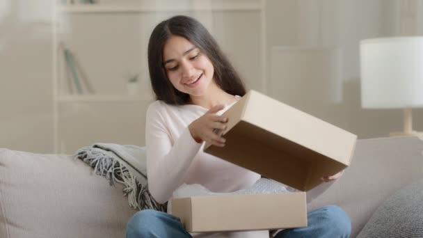 Amazed arabian latino female customer shopper buyer sitting on sofa unpacking postal shipping delivery parcel box gift present surprised woman girl enjoying internet order purchases from online store - Imágenes, Vídeo