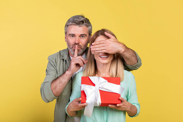 Mature man got surprise for his loving wife, woman holding gift box, man covering her eyes with palm and showing silence gesture, yellow studio background - Foto, Imagen