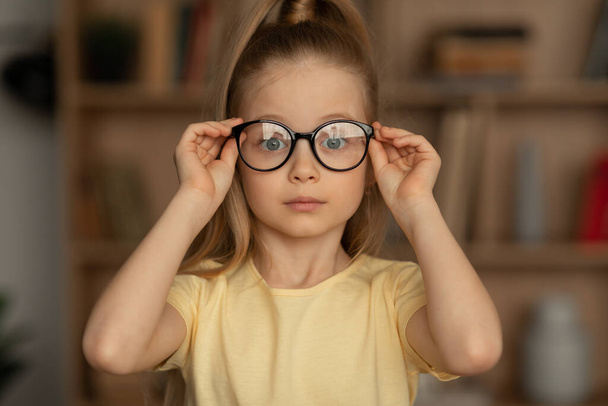 Zicht. Portret van een klein meisje met een bril die naar de camera kijkt terwijl ze poseert in de bibliotheek. Kinderen ogen gezondheid en zicht Correctie Concept. Vooraanzicht - Foto, afbeelding