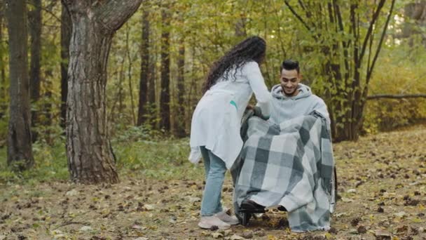 Young nurse in medical gown covers guy with warm blanket takes care patient in wheelchair caring girl visits friend in rehab walks in autumn hospital park enjoy walking outdoors rehabilitation center - Imágenes, Vídeo