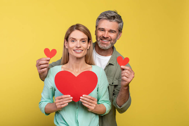 Love is in the air. Loving middle aged spouses with red paper hearts in hands posing over yellow studio background. Happy man and woman smiling at camera - Photo, Image