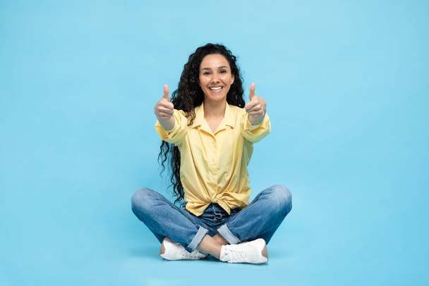 I Like It. Cheerful Arabic Woman Gesturing Thumbs Up With Both Hands Sitting Posing Over Blue Background. Female Approving Offer Smiling To Camera. Studio Shot - 写真・画像
