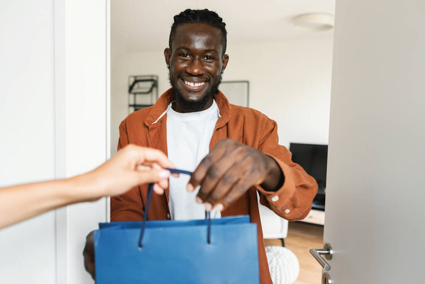 Shopping delivery service. Happy african american man getting shopper bags from courier and smiling at camera, black guy standing near door of his home. - Zdjęcie, obraz