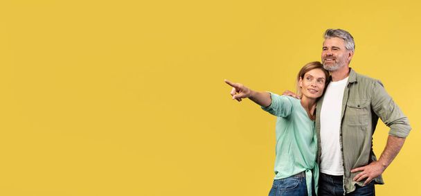 Check this great offer. Positive middle aged spouses hugging and woman pointing aside at copy space, demonstrating empty place for advert mockup, yellow studio background, panorama - Foto, Bild