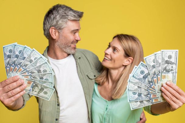 Big luck and win concept. Happy mature spouses holding a lot of dollar cash, celebrating success together, posing with money over yellow studio background - Fotó, kép