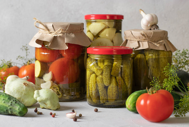 Pickled cucumbers, tomatoes and squash with dill, garlic and chili peppers in glass jars on light gray background - Photo, image