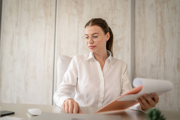 One woman female caucasian entrepreneur businesswoman or secretary sitting at her office at desk work checking paper documents contract wear white shirt copy space - Foto, imagen