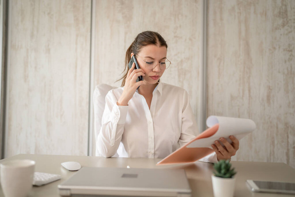 One woman female caucasian entrepreneur businesswoman or secretary sitting at her office at desk work happy smile making a phone call talk checking paper documents contract wear white shirt copy space - Fotografie, Obrázek