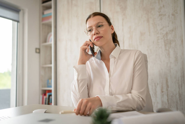One woman female caucasian entrepreneur businesswoman or secretary sitting at her office at desk work happy smile making a phone call talk checking paper documents contract wear white shirt copy space - Foto, Imagen