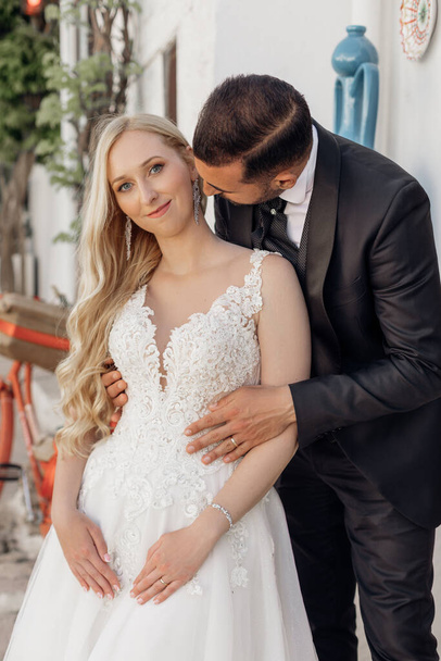 Portrait of beautiful wedding couple standing on street. Young woman with long wavy fair hair wearing long white embroidered wedding dress, looking at camera, groom in suit hugging bride from behind.  - Photo, Image