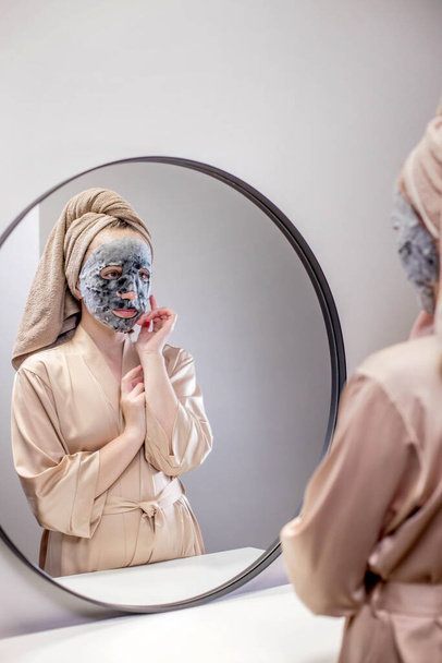 Young happy woman in towel making face mask and looking in mirror in stylish bathroom. The woman uses a mask to cleanse the skin. Skin care. - Foto, Bild
