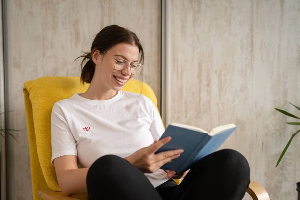 One woman young adult caucasian female sitting in chair at home read book copy space front view real people leisure weekend concept looking to the book happy smile - Foto, Bild