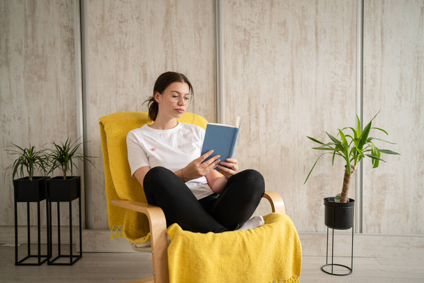Een vrouw jong volwassen blanke vrouw zitten in stoel thuis lees boek kopiëren ruimte vooraanzicht echte mensen vrijetijdsweekend concept - Foto, afbeelding