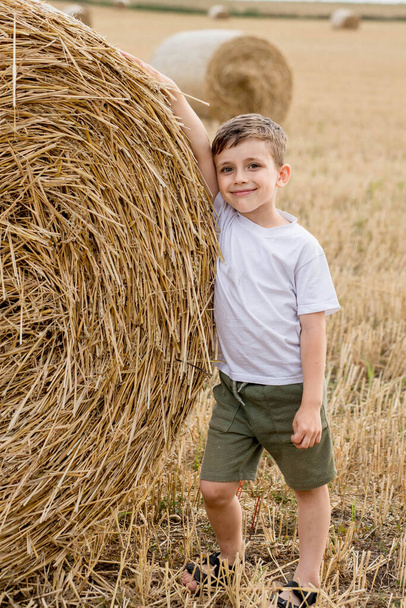 A cute little boy in a white t-shirt and khaki shorts is standing near round bales of hay. Photo session in the field. - Foto, afbeelding