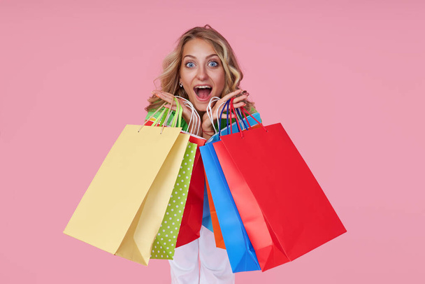 Portrait of an excited joyful young woman with colorful shopping bags on a pink background. Surprised Girl with wide open mouth - Фото, изображение