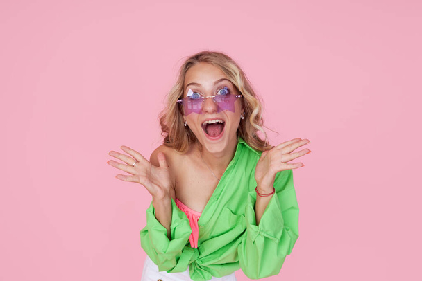 Portrait of pretty cheerful girl with wide open mouth in green bright shirt and star shape sunglasses isolated on pink background - Fotó, kép
