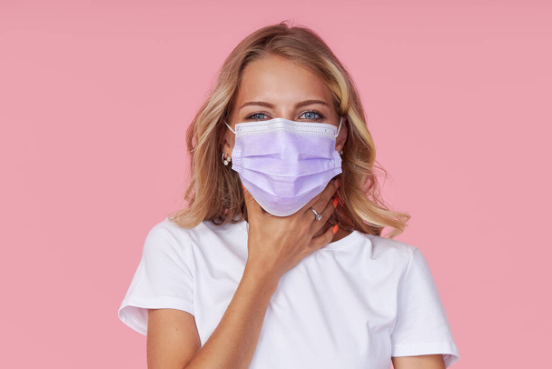 Young ill woman in a white casual t-shirt wears a protective face mask. holding hands on neck. Protection against flu and cold diseases during a pandemic. Pink isolated background - Foto, Bild