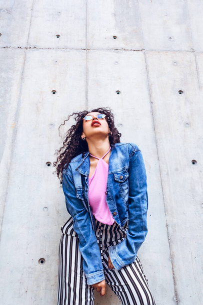 beautiful teen woman with glasses and curly hair wearing trendy blue modern jacket on gray city wall posing, shot from below, generation z, urban concept. - Фото, изображение