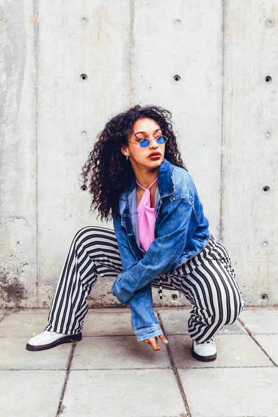 Curly-haired teen woman in blue glasses sitting in stylish urban fashion pose. Generation z woman posing in the city with glasses - Zdjęcie, obraz