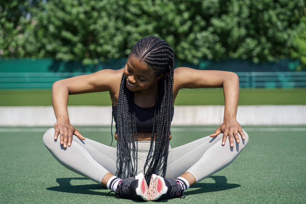 Молодая афроамериканка с длинными темными косичками любит растягивать ноги перед интенсивной тренировкой. Активная самка сидит на зеленой дорожке на спортивной площадке против пышных зеленых деревьев в солнечный летний день - Фото, изображение