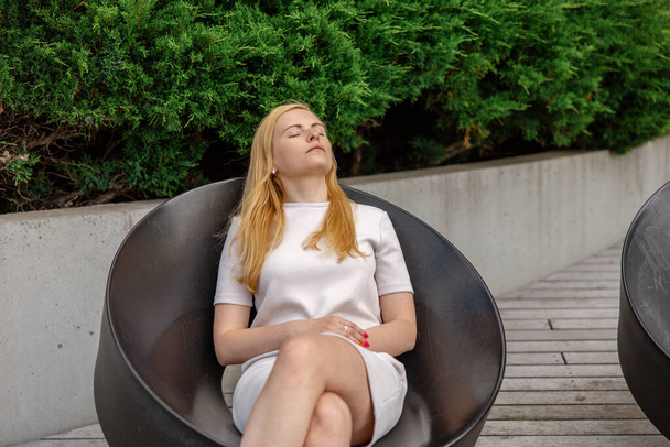 Young beautiful blond woman sitting outdoors, on the wooden terrace in the city and resting. Girl has break, spending time outside, relaxing. Time with yourself, dreaming, relaxation, mental health - Fotografie, Obrázek