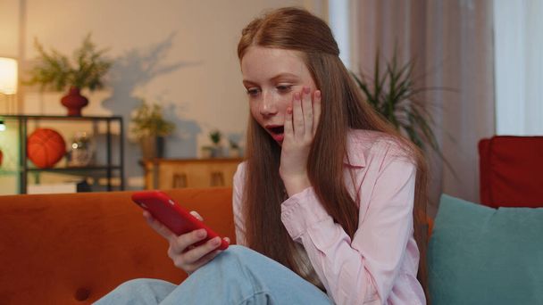 Oh my God, Wow. Happy excited teen young child girl use mobile smartphone typing browsing say yes found out great big jackpot lottery win, good news, celebrate at home couch. Ginger children kid alone - Foto, imagen