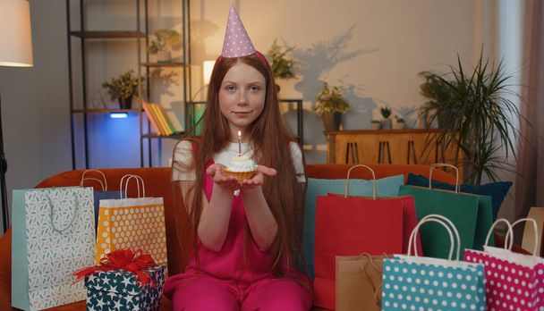 Happy teen child girl wears festive birthday hat hold cupcake makes wish joyful congratulating blowing burning candle on cake. Ginger kid celebrating anniversary party on couch with gifts at home - Photo, Image