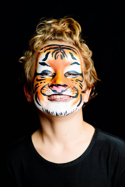 Πορτρέτο ενός αγοριού με ζωγραφισμένο πρόσωπο τίγρης με χαρούμενη έκφραση, απομονωμένο σε μαύρο φόντο. - Φωτογραφία, εικόνα