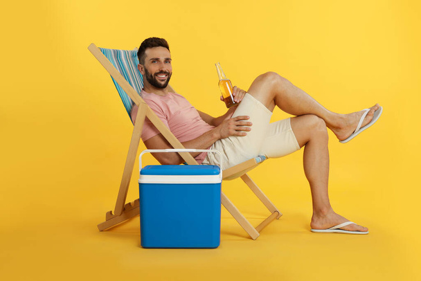 Happy man with bottle of beer resting in deck chair near cool box on yellow background - Foto, Imagem