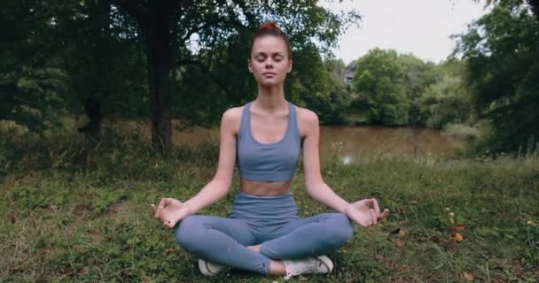 Mujer meditando en pose de loto mientras realiza un bajo yoga asana al aire libre en un parque en ropa deportiva sobre hierba verde en el verano. Imágenes de alta calidad 4k - Metraje, vídeo