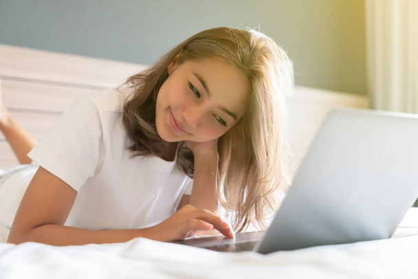 Belle femme asiatique couchée et souriant le lit et son ordinateur portable. WFH Travailler de la maison en quarantaine. Éloignement social Auto-isolement - Photo, image