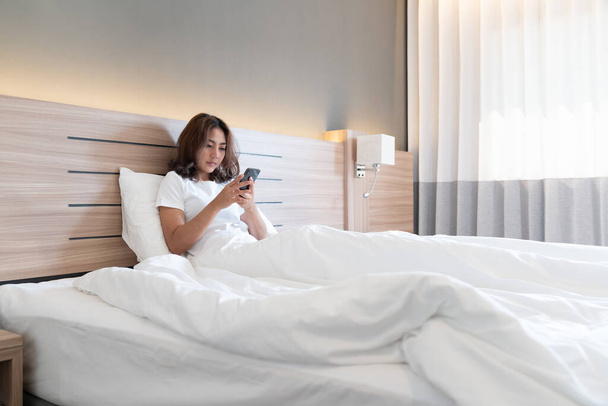 Νεαρές Ασιάτισσες ξαπλωμένες στο κρεβάτι στέλνοντας μηνύματα και τσεκάροντας κοινωνικές εφαρμογές σε smartphone στο κρεβάτι πριν κοιμηθεί το βράδυ. Έννοια κινητού εξαρτημένου. - Φωτογραφία, εικόνα