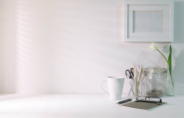 Comfortable workplace. Blank picture frame, coffee cup, notebook and stationery on white table with sunlight through blinds by the window. - Foto, Bild