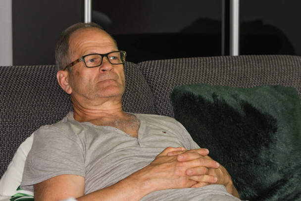  Biały starszy mężczyzna z siwymi włosami w luźnej koszulce i okularach oglądający wiadomości w telewizji i śpiący na kanapie. Wieczorna rozrywka w domu. Pojęcie życia w podeszłym wieku. - Zdjęcie, obraz