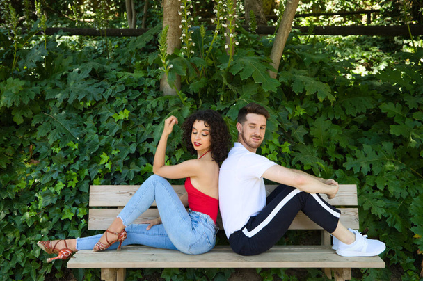 Egy jóképű fiatalember és egy gyönyörű nő ülnek a fa padon háttal egymásnak. Mindegyik más-más oldalra néz, ahogy boldogan néznek egymásra a szemük sarkából.. - Fotó, kép