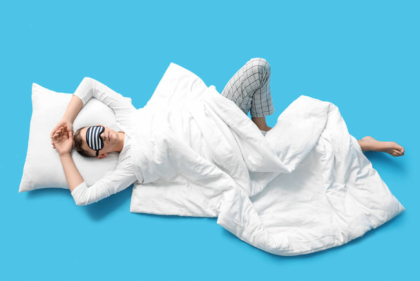 Young man with sleeping mask, pillow and blanket lying on blue background - Zdjęcie, obraz