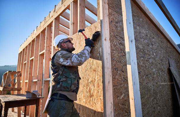 Carpenter hammering nail into OSB panel on the wall of future cottage. Man worker building wooden frame house. Carpentry and construction concept. - Foto, afbeelding