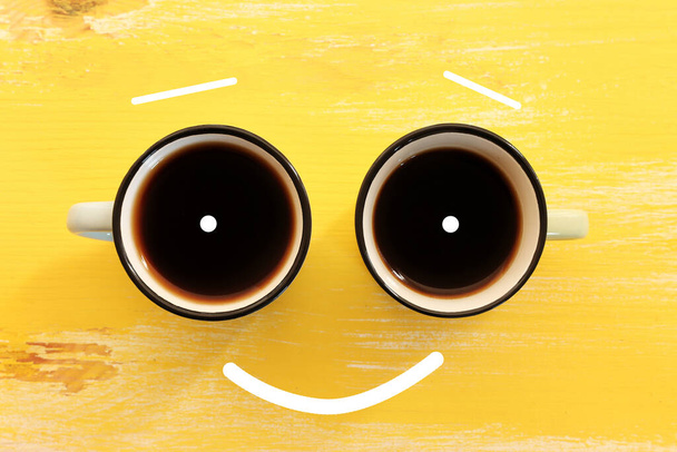 Top view image of coffee cups and smiling face over yellow wooden table. Happiness and joy concept - Φωτογραφία, εικόνα