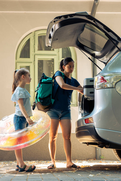 Woman with little girl going on holiday with automobile, travelling to seaside with luggage and inflatable. Leaving on summer vacation, loading travel bags and trolley in vehicle trunk. - Foto, Imagen