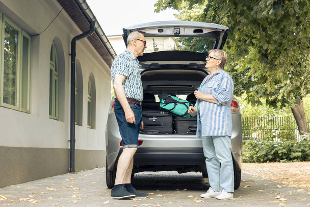 Elderly couple going on holiday vacation, loading suitcase and luggage in automobile trunk at home. Travelling by car with baggage and bags, leaving onubran cityscape adventure trip. - Foto, afbeelding