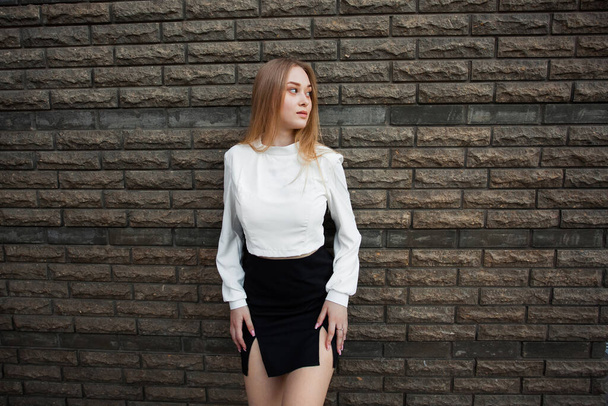 A slender young schoolgirl with blond hair, in a white shirt and a short black skirt, stands against a dark textured stone wall, looks away and keeps her hands on her hips. - Photo, image
