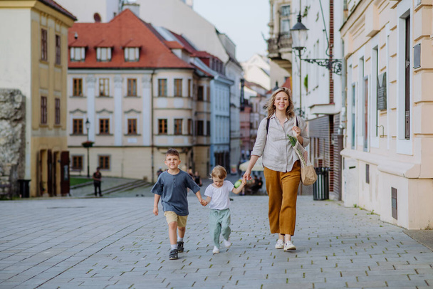 A young mother with zero waste shopping bag holding hands with her children and walking in old city street center. - 写真・画像