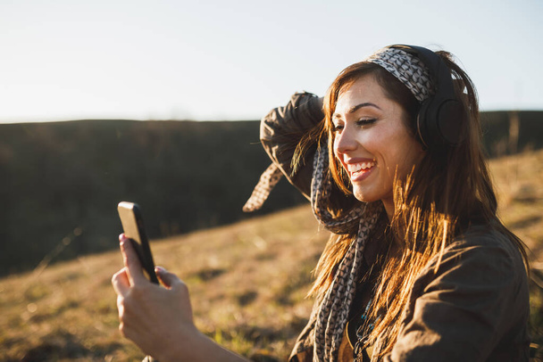 Smiling young woman making video call by smartphone while sitting on the ground in sunny day and listening music. - Foto, Imagen