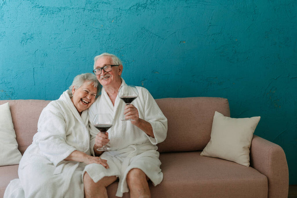 Happy senior couple sitting together in the bathrobe on sofa with glass of wine, having nice time at home. - Фото, зображення